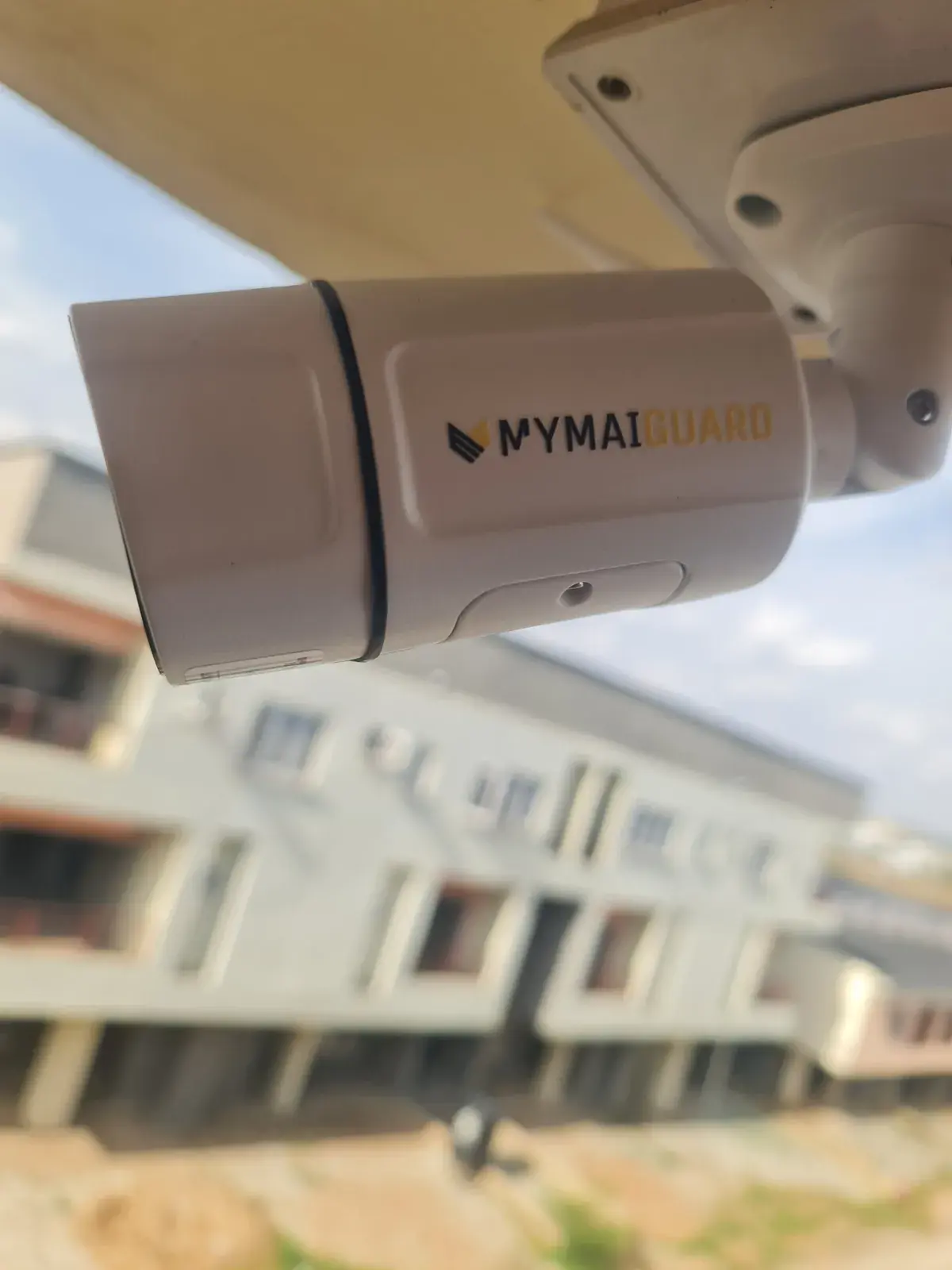 Close-up of a MYMAiGUARD-branded outdoor bullet camera fixed under a roof edge, with houses in the background